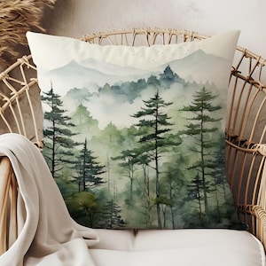 May include: Square cushion with a watercolour painting of a misty forest scene. The design shows tall evergreen trees in shades of green and grey, with mountains in the background. The cushion is placed on a wicker chair.