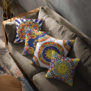 Może przedstawiać: Cztery dekoracyjne poduszki z kolorowymi, ozdobnymi wzorami leżą na szarej sofie. Poduszki mają wzory słoneczne, kwiatowe i geometryczne w odcieniach niebieskiego, żółtego, czerwonego i białego. Sofa jest wykonana z tkaniny w neutralnym kolorze.