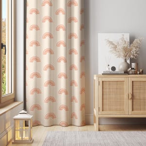 Puede incluir: Cortina color crema con un patrón repetido de arcoíris color melocotón. La cortina cuelga frente a una ventana, y en el fondo se ve un mueble de madera con frente tejido. Una linterna decorativa está en el suelo.