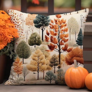 May include: A cushion featuring a forest design with trees in autumnal colours of orange, green, and yellow. The cushion is placed next to a pot of orange flowers and two pumpkins. The background of the cushion is cream-coloured.