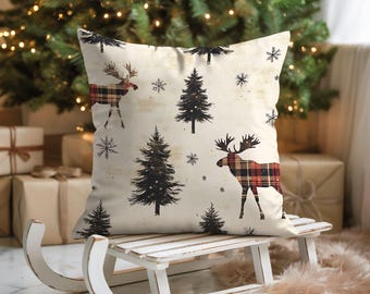 May include: A cream-coloured cushion with a festive Christmas design. The pattern includes black fir trees, snowflakes, and moose with red and black plaid patterns. The cushion is placed on a white wooden sled, evoking a winter scene.