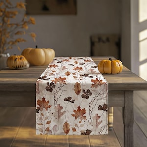 May include: A fall-themed table runner with a cream background and a repeating pattern of brown and orange leaves. The runner is displayed on a wooden table, with pumpkins in the background, creating a seasonal autumn aesthetic.