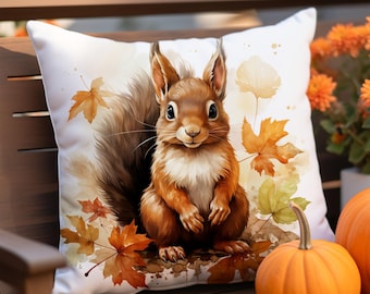Könnte beinhalten: Quadratisches weißes Kissen mit einer Aquarellillustration eines Eichhörnchens und Herbstblättern. Das Eichhörnchen ist braun und weiß mit einem flauschigen Schwanz. Das Kissen ist mit orangefarbenen und gelben Ahornblättern verziert, ideal für die Herbstdekoration.