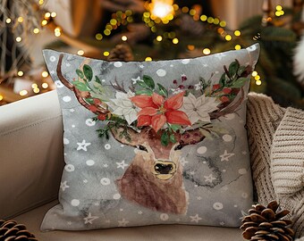Puede incluir: Un cojín cuadrado decorativo con una ilustración de acuarela de una cabeza de ciervo adornada con una corona floral. El cojín tiene un fondo gris con copos de nieve blancos y detalles de estrellas. El ciervo tiene cuernos marrones y una flor de pascua roja.