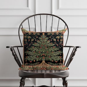 Può includere: Un cuscino quadrato decorativo con un design dettagliato di albero di Natale. L'albero è verde con bacche rosse e ornamenti dorati, su sfondo nero. Il cuscino ha un bordo beige ed è posto su una sedia di legno scuro.