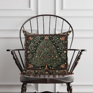 May include: A decorative square pillow with a dark green background and a detailed Christmas tree design. The tree is adorned with red ornaments and gold accents, set against a floral border. The pillow is placed on a dark wooden chair.