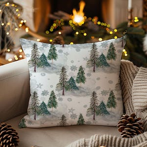 Op de afbeelding: Een vierkant sierkussen met een winterbosontwerp. Het patroon bevat groenblijvende bomen, sneeuwvlokken en een lichtgrijze achtergrond. Het kussen ligt op een beige bank, met een warme deken en dennenappels. De scène roept een gezellige sfeer op.
