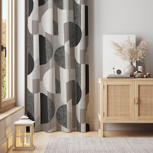May include: A geometric patterned curtain in shades of grey, black, and beige. The curtain hangs in front of a window. A wooden cabinet with a woven front is visible to the right. A small lantern sits on the windowsill.