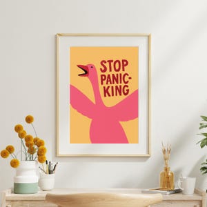 May include: Framed art print with a pink goose and the text "STOP PANICKING" in red against a yellow backdrop. The artwork is in a wooden frame, ideal for wall decor. The image is a fun and quirky piece.