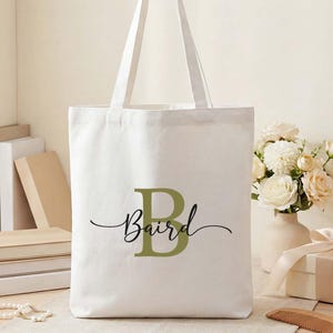 May include: A white canvas tote bag with the name "Baird" printed in a script font, with a large green letter "B". The bag is on a wooden table with books, flowers, and a gift box.