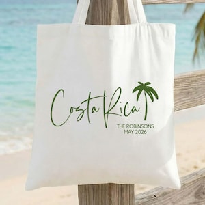 May include: White tote bag with the words "Costa Rica" in a script font and a palm tree graphic in green. The text "THE ROBINSONS MAY 2026" is below. The bag is hanging on a wooden fence with a beach and ocean in the background.