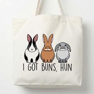 May include: Cream-colored tote bag featuring three cartoon rabbits in black and white, brown, and gray. The text "I GOT BUNS, HUN" is printed below the rabbits in black. The bag has two handles.