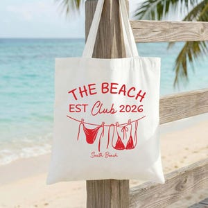 Puede incluir: Un bolso de tela blanco con texto en rojo que dice THE BEACH EST Club 2026 y South Beach. Debajo del texto, un gráfico rojo muestra un bikini y una parte superior de traje de baño colgados de un tendedero con pequeñas pinzas.