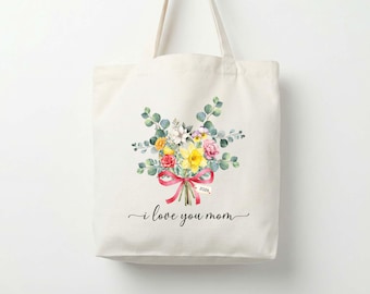 Puede incluir: Un bolso de tela color crema con una ilustración en acuarela de un ramo de flores con narcisos amarillos, rosas rosadas y hojas de eucalipto verdes atadas con una cinta roja. Una pequeña etiqueta dice 2026. El texto I love you mom aparece debajo.