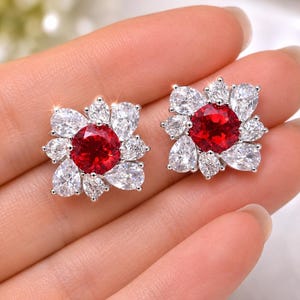May include: A pair of flower-shaped stud earrings. Each earring features a large, round, red gemstone at the center, surrounded by white, pear-shaped stones. The earrings are set in a silver-colored metal and are displayed against a neutral background.