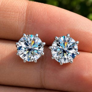 May include: A pair of round-cut stud earrings featuring clear, faceted stones set in six-prong silver-toned metal settings. The stones exhibit high brilliance and light refraction, creating a sparkling appearance with distinct geometric facets.