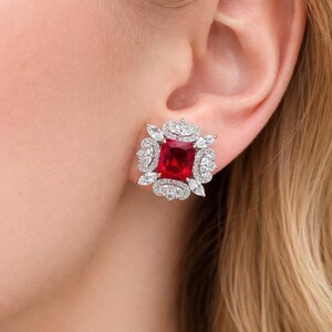 May include: A square-shaped stud earring with a large, vibrant red center stone. The red stone is surrounded by smaller, clear, round and marquise-cut stones set in a silver-colored metal. The earring is shown on a person's ear.