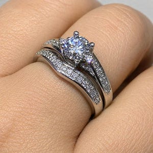 Peut inclure: Une bague de fiançailles argentée avec une grande pierre centrale ronde taille brillant fixée par quatre griffes. L'anneau et l'alliance incurvée assortie sont tous deux incrustés de nombreuses petites pierres serties pavé.