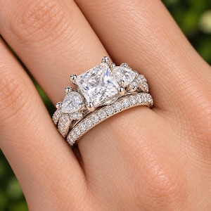 May include: A silver-toned engagement ring set featuring a central princess-cut stone flanked by two pear-shaped stones, with a matching band encrusted with small round stones along the entire visible surface of both rings.