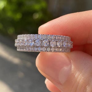 May include: A silver-toned band ring featuring a central row of large, round, clear stones flanked by two outer rows of smaller, pavé-set clear stones. The metal surface is highly reflective, creating a bright, sparkling appearance across the entire ring.
