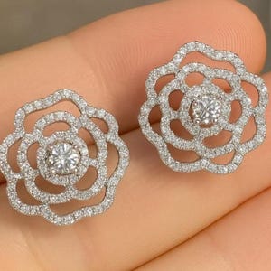 May include: A pair of silver-toned stud earrings shaped like open-work flowers. Each earring features a central round brilliant-cut stone surrounded by concentric layers of smaller pavé-set stones, creating a delicate, layered floral silhouette.