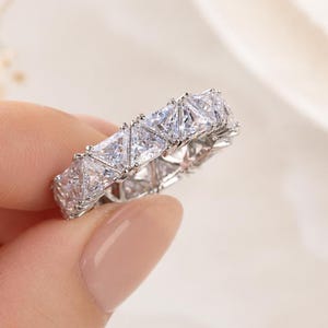 May include: A silver-toned eternity band ring featuring a continuous row of clear, triangular-cut stones set in a metallic frame. The stones are arranged in a repeating pattern around the entire circumference of the circular jewellery piece.