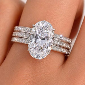 May include: A three-piece diamond ring set. The engagement ring features a large oval-cut diamond. Two additional bands are encrusted with smaller diamonds. The rings are set in a silver-coloured metal.