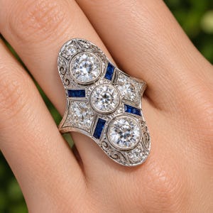 May include: An elongated silver-toned ring featuring three prominent round-cut clear stones arranged vertically, accented by smaller clear stones and rectangular deep blue sapphires, all set within an intricate metal frame with scrollwork details.