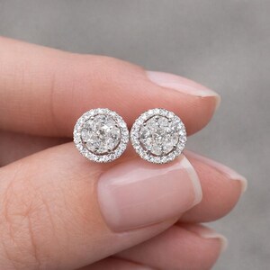May include: A pair of diamond stud earrings. Each earring features a cluster of diamonds surrounded by a halo of smaller diamonds, set in a silver or white gold setting. The earrings are round and sparkling.