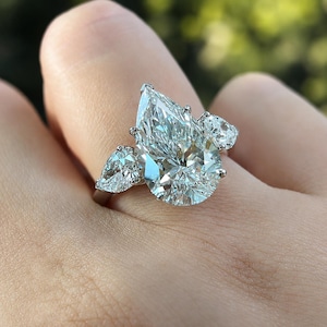 Könnte beinhalten: Ein Ring mit drei Steinen, bestehend aus einem großen birnenförmigen Diamanten in der Mitte, der von zwei kleineren birnenförmigen Diamanten flankiert wird. Die Steine sind in ein poliertes, silberfarbenes Metallband gefasst und glänzen.