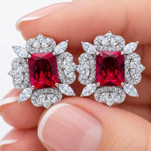 May include: A pair of ornate stud earrings featuring a vibrant red rectangular center stone surrounded by smaller, sparkling white stones. The earrings have a floral-like design with a crown-shaped top, creating a luxurious and elegant look.