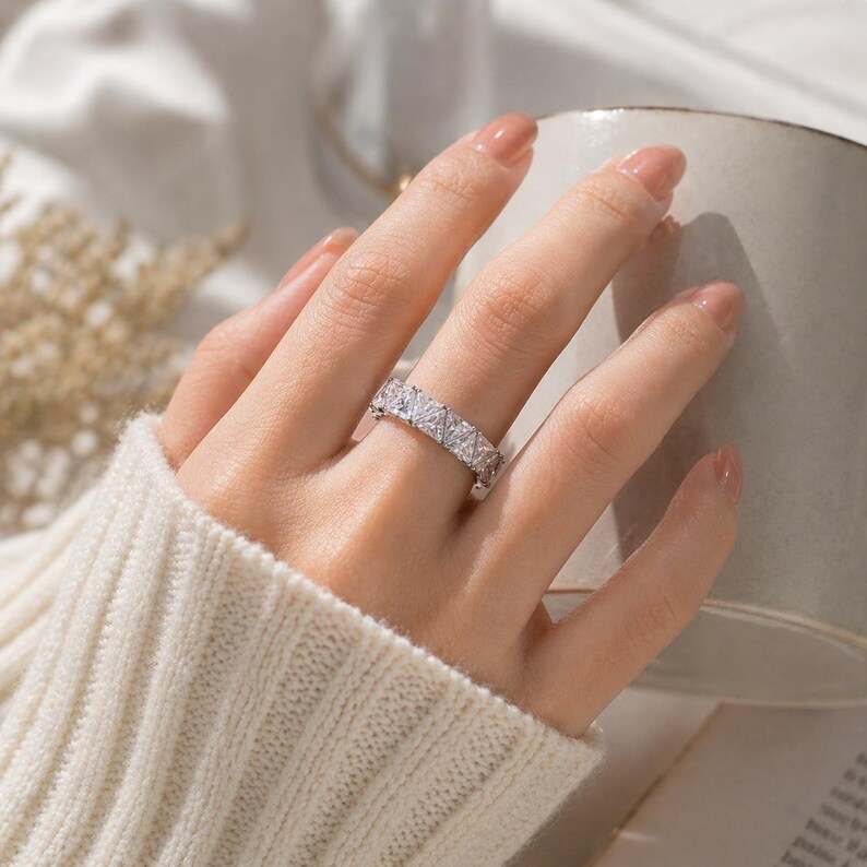 Puede incluir: Un anillo plateado con un patr&oacute;n repetitivo de piedras rectangulares y transparentes. El anillo se lleva en una mano con u&ntilde;as cuidadas, sobre un fondo suave y neutro.