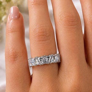 Puede incluir: Un anillo plateado con una fila de piedras preciosas claras, de corte cuadrado. El anillo se lleva en un dedo, con la mano enfocada. Las piedras est&aacute;n engastadas juntas, creando un brillo continuo. Un dise&ntilde;o cl&aacute;sico, adecuado para diversas ocasiones.