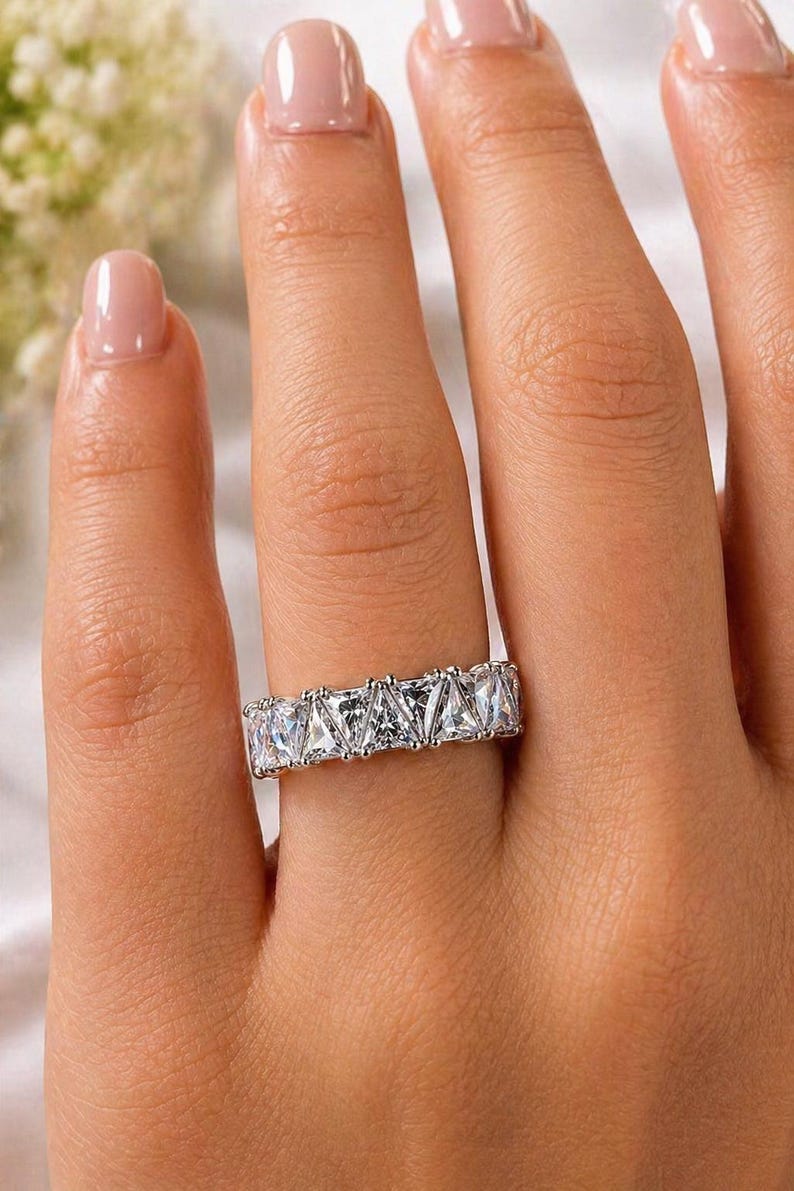 Puede incluir: Un anillo plateado con una fila de piedras preciosas claras, de corte cuadrado. El anillo se lleva en un dedo, con la mano enfocada. Las piedras est&aacute;n engastadas juntas, creando un brillo continuo. Un dise&ntilde;o cl&aacute;sico, adecuado para diversas ocasiones.