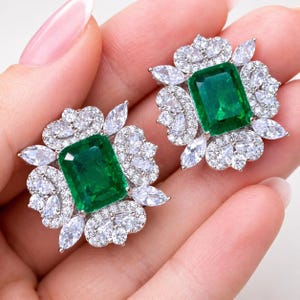 May include: A pair of stud earrings featuring a large, rectangular, emerald-green center stone surrounded by a cluster of clear, marquise-cut and round-cut white stones set in a silver-toned metal frame with a floral-inspired arrangement.