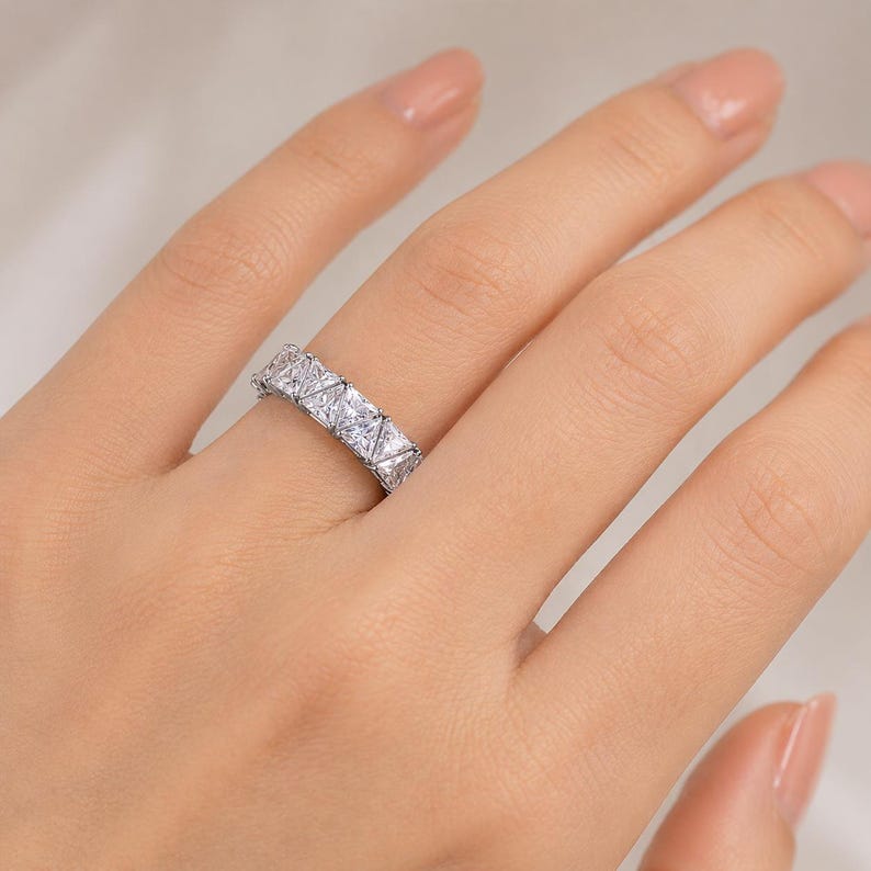 Puede incluir: Un anillo plateado con una serie de piedras preciosas claras de corte cuadrado. El anillo est&aacute; en un dedo, mostrando el intrincado dise&ntilde;o y el brillo de las piedras. Una joya.