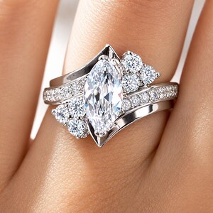 May include: A silver-toned ring featuring a large marquise-cut center stone flanked by clusters of small round stones. The band is split into two curved sections, each accented with a row of tiny pavé-set stones that follow the contour of the ring.
