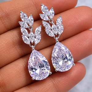 May include: A pair of silver-toned drop earrings featuring a cluster of marquise-cut clear stones arranged in a leaf-like pattern at the top, connected to a large, faceted pear-shaped clear stone pendant that hangs vertically from the base.