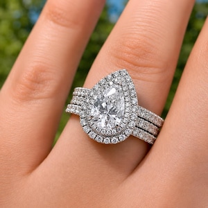 Peut inclure: Une bague argentée avec une pierre centrale en forme de poire entourée d'un double halo de petites pierres rondes. L'anneau est divisé en trois rangées distinctes, chacune incrustée de petites pierres rondes, créant un aspect large et superposé.