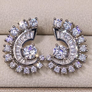 May include: A pair of silver-toned earrings featuring a crescent-shaped design. Each earring is adorned with a central round-cut stone, a curved row of baguette-cut stones, and a border of small round-cut stones along the outer edge.