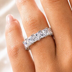 Puede incluir: Un anillo plateado con una serie de piedras preciosas claras, de corte cuadrado. El anillo est&aacute; en un dedo, y las piedras preciosas reflejan la luz, creando un efecto brillante. El anillo es una joya.
