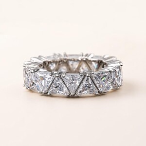 Puede incluir: Un anillo plateado con una banda continua de piedras transparentes de corte triangular. La montura del anillo tambi&eacute;n es plateada, creando un dise&ntilde;o brillante y sin costuras. El anillo es de estilo eterno cl&aacute;sico.
