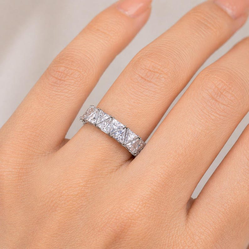 Puede incluir: Un anillo plateado con una fila de piedras preciosas claras, talladas en forma cuadrada. El anillo est&aacute; en un dedo, con la mano y el fondo ligeramente borrosos.