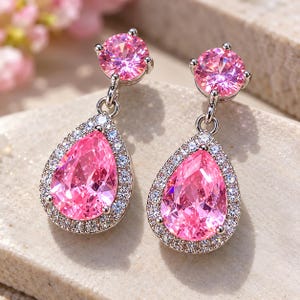 May include: A pair of drop earrings featuring a round pink stone at the top and a larger pear-shaped pink stone at the bottom, both surrounded by a halo of small clear stones set in a silver-toned metal frame with a secure post backing.