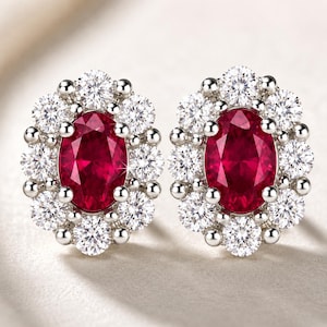 Peut inclure: Une paire de boucles d'oreilles à clous avec une pierre précieuse centrale rouge foncé de taille ovale, entourée d'un halo de douze petites pierres blanches rondes et brillantes, serties dans une monture en métal argenté poli.