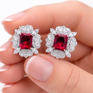 May include: A pair of square-cut, ruby-red gemstone stud earrings. Each earring is surrounded by a halo of small, clear, round and marquise-cut gemstones, set in a silver-colored metal. The earrings are held in a person's hand.