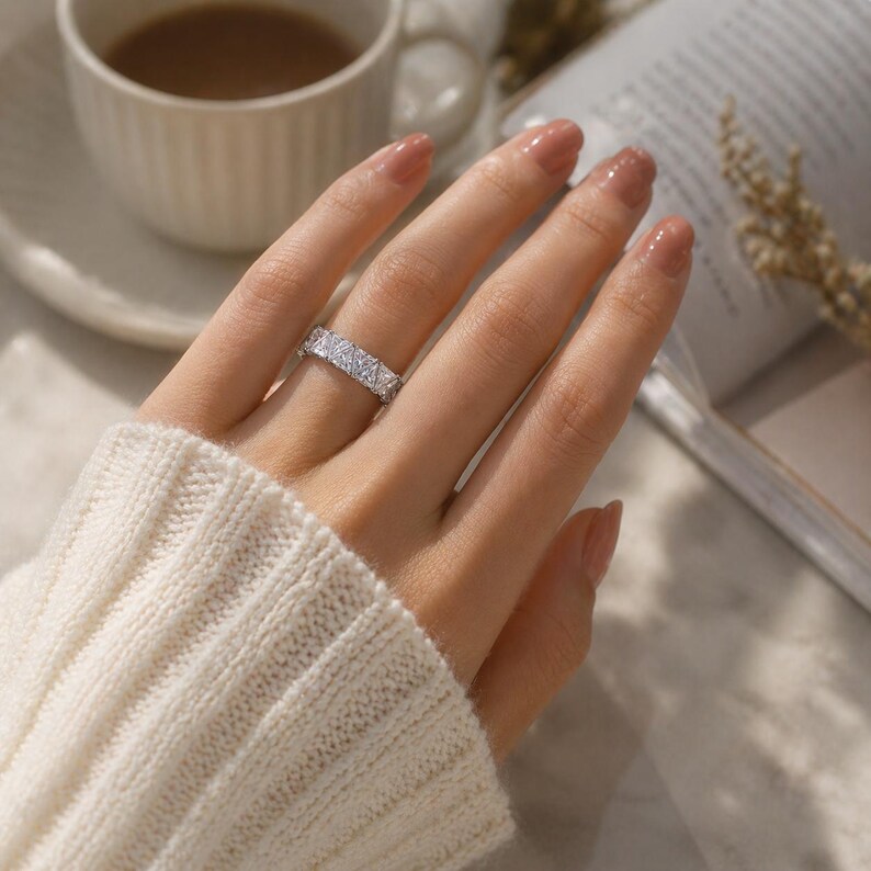 Puede incluir: Un anillo plateado con un dise&ntilde;o geom&eacute;trico con m&uacute;ltiples piedras peque&ntilde;as de corte cuadrado. El anillo se lleva en un dedo, con un su&eacute;ter blanco en el fondo. El anillo es una joya.