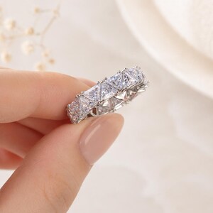 Puede incluir: Un anillo plateado con una banda continua de piedras preciosas claras, talladas en forma cuadrada. El anillo se muestra sobre un fondo suave y neutro, resaltando sus facetas brillantes. El dise&ntilde;o del anillo es de estilo eterno cl&aacute;sico.