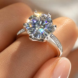May include: A silver-toned ring featuring a large, round, brilliant-cut clear stone held by six prongs. The band is accented with small, pavé-set clear stones along the shoulders, creating a reflective, sparkling appearance in the light.