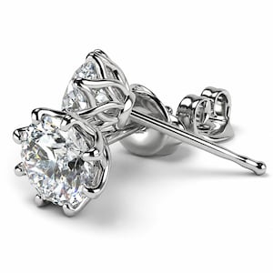 May include: A pair of silver-toned stud earrings featuring round, clear, faceted stones set in a six-prong floral-inspired metal setting. Each earring includes a matching silver-toned post and a secure butterfly-style friction back closure.